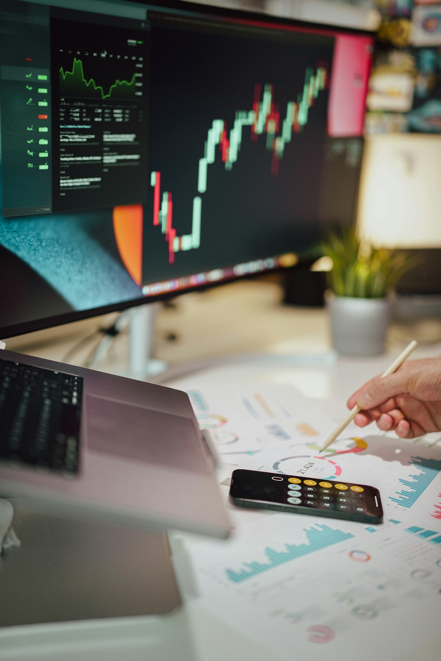 Person arbeitet an einem Schreibtisch mit einem Laptop, Smartphone und gedruckten Diagrammen. Auf dem grossen Monitor ist ein Börsendiagramm mit steigenden und fallenden Kurven zu sehen. Die Person zeigt mit einem Stift auf ein Diagramm auf dem Tisch, neben dem Smartphone, das einen Taschenrechner anzeigt.