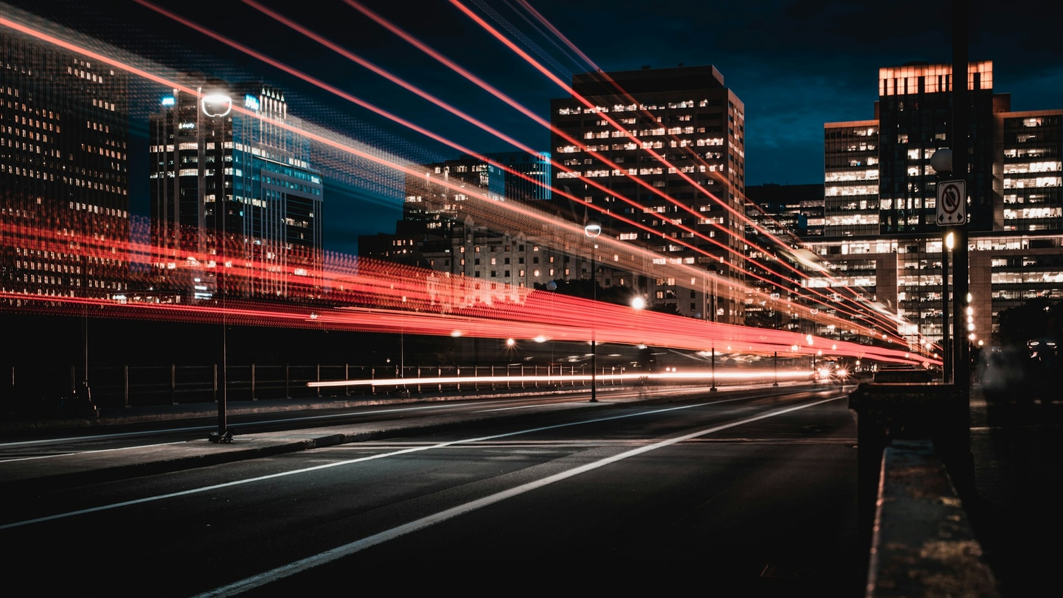 Stadtansicht bei Nacht mit Hochhäusern im Hintergrund. Über eine Strasse ziehen sich lange Lichtspuren von Autos, die rote und weisse Linien in Bewegung hinterlassen, was auf eine Langzeitbelichtung hindeutet.